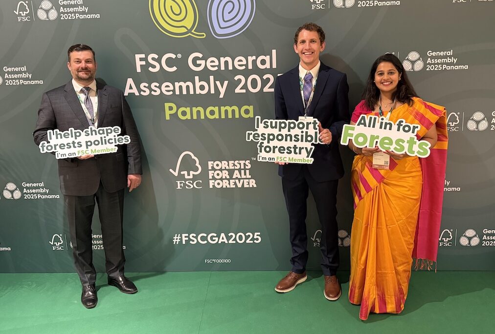 Chris, Nathan and Anusha are posing in front of the FSC General Assembly 2025 backdrop and holding inspirational signs.