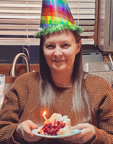 Jess wearing a rainbow-colored birthday hat and holding a piece of birthday cake with a lit candle in it