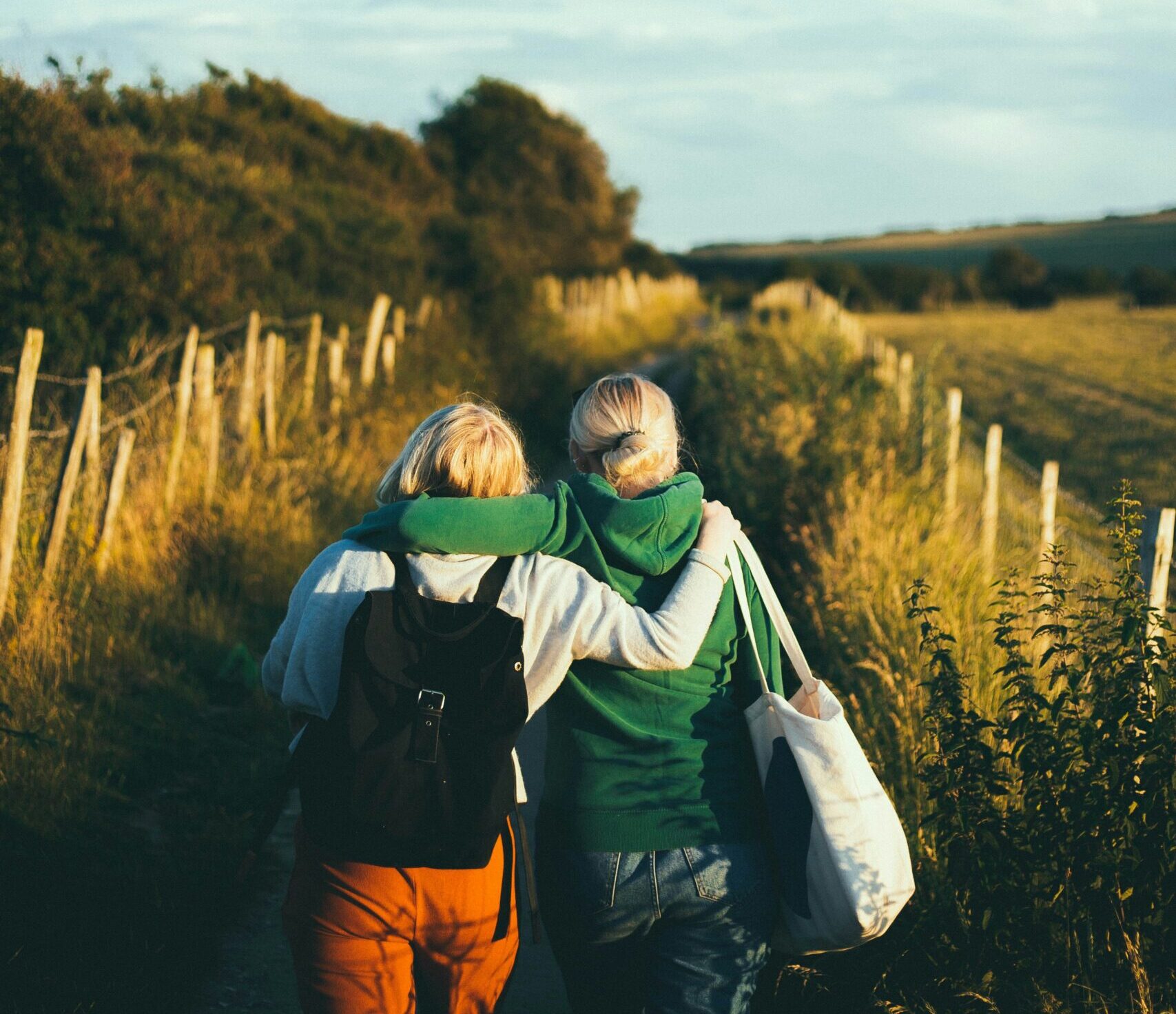 Two friends, one with her arm around the other, are walking along a wooded road towards a beautiful blue sky.