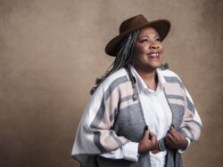 Melody Mobley poses against and brown background. She's smiling and wearing brown Western-style hat and a gray and white shawl.