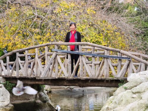 Sarah is standing the middle of a wooden bridge above reflective, flat water.