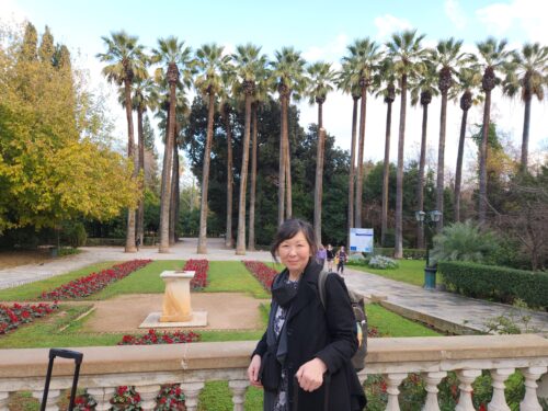 Sarah is smiling and posing for the camera in front of a beautiful garden of red flowers and palm trees