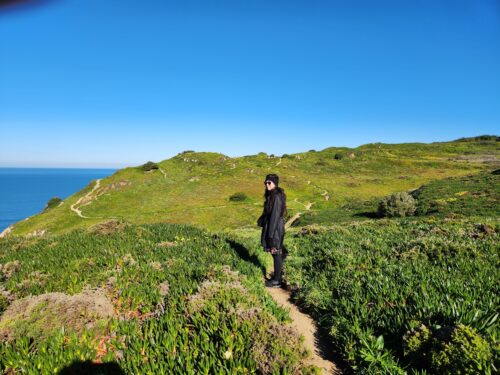 Sarah is bundled up on a chilly, blue-sky day. She's looking at the camera from a dirt path overlooking the ocean.